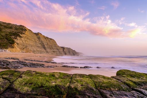A beach surrounded by the sea and cliffs covered in mosses under a cloudy sky during the sunset
