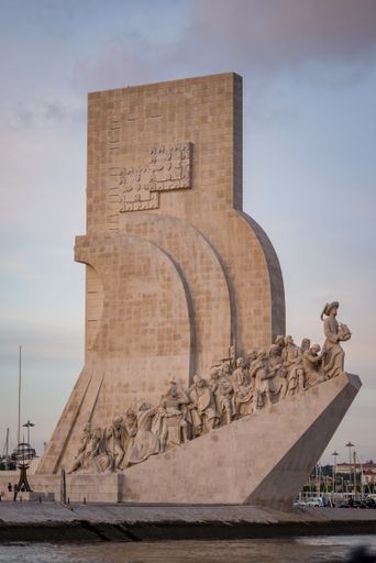 The Monument of the Discoveries surrounded by buildings and water under a cloudy sky in the evening