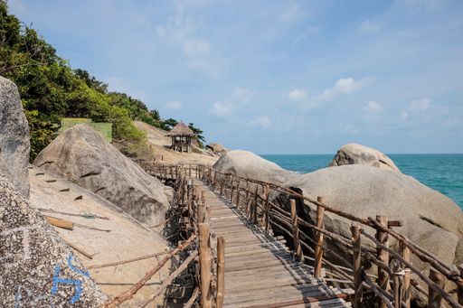 A wooden bridge surrounded by rocks covered in greenery and the sea under the sunlight