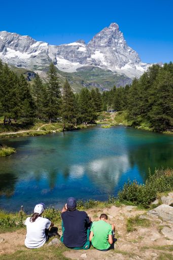 A vertical shot of a people sitting near a pond surrounded by trees with a mountain in the distance in Cervinia Breuil-Cervinia, Italy