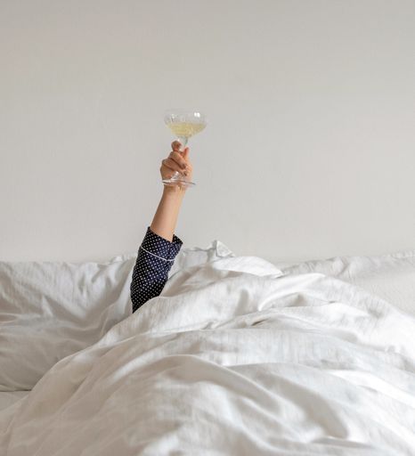 Person in pajamas covered with bed linen, arms raised, holding and glass., wi Celebration, celebrating, new year, cozy, lifestyle, home.