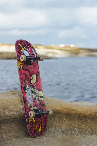 A closeup shot of a pink skateboard leaning on a rock next to a beach
