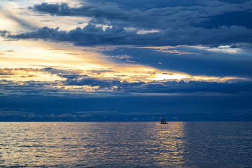 A panoramic shot of a sea with a lone sailboat under  cloudy blue skies with the sun hiding behind the clouds