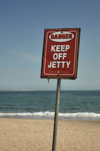 A red sign at the sandy beach that reads "DANGER KEEP OFF JETTY" with the sea and beautiful clear sky