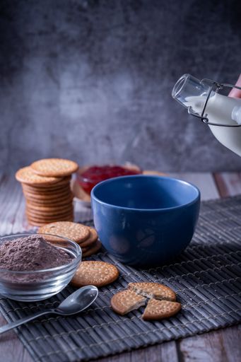 A vertical shot of milk in a bottle, a bowl next to cocoa powder, Maria cookies, and jam