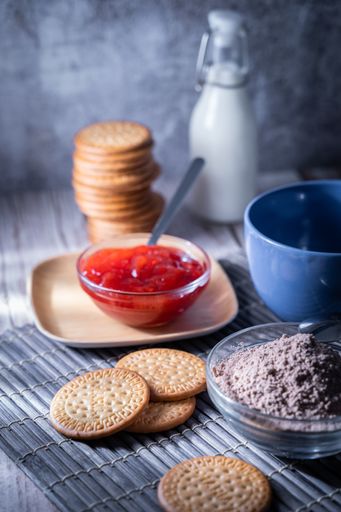 A vertical shot of fresh Maria cookies, strawberry jam, cocoa powder, a bottle omilk, and a bowl