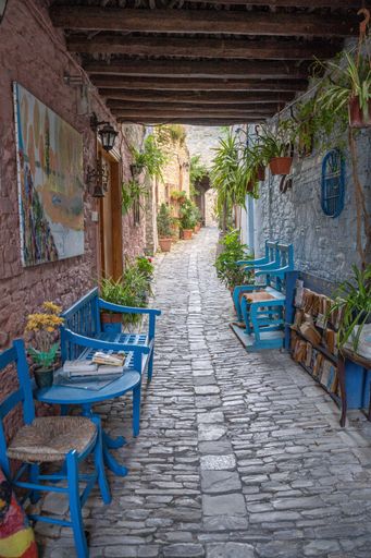 A vertical shot of a narrow bright tunnel in Lefkara, Cyprus with plants and lamps on the walls along with blue chairs on cobbled alley
