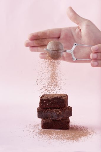 A closeup shot of hands powdering cacao powder on a piece of freshly baked chocolate brownie cake