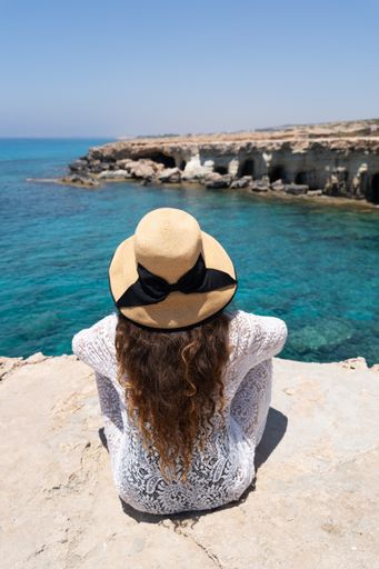 A back view of a woman in a white summer dress and a beautiful hat sitting on a cliff on the coast