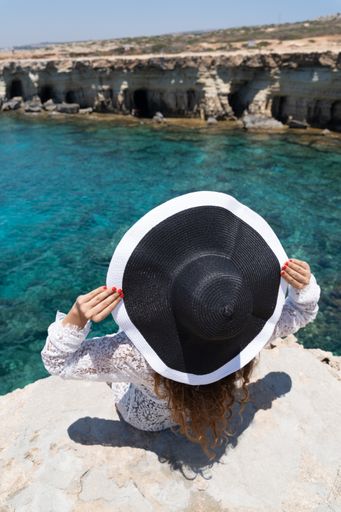 A woman with a big black hat leaning back on a beach, looking at the sea caves in Ayia Napa, Cyprus