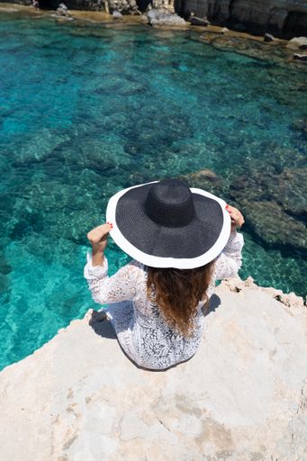 A back view of an elegant woman sitting on a cliff wearing a black wide brimmed hat and a summer dress