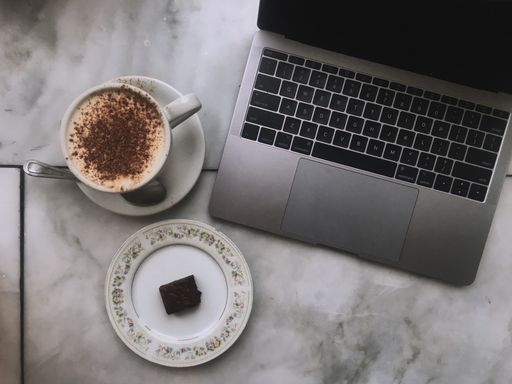 An overhead shot of a laptop with a cappuccino and a brownie on a ceramic surface