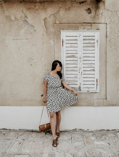 A full body shot of a young sexy woman in a light polka dot dress standing against an old cracked white wall holding her skirt