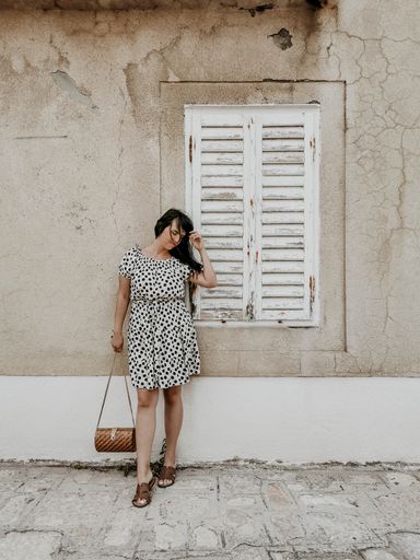 A full body shot of a young sexy smiling woman in a light polka dot dress standing against an old cracked white wall fixing her hair