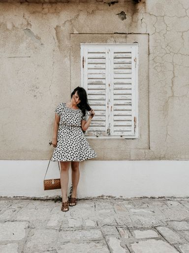 A full body shot of a young sexy smiling woman in a light polka dot dress standing against an old cracked white wall fixing her hair