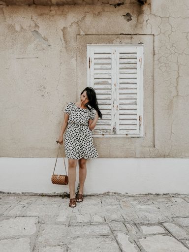 A full body shot of a young sexy smiling woman in a light polka dot dress standing against an old cracked white wall fixing her hair