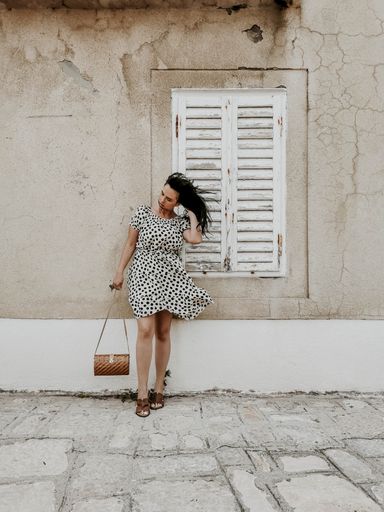 A full body shot of a young sexy smiling woman in a light polka dot dress standing against an old cracked white wall fixing her hair