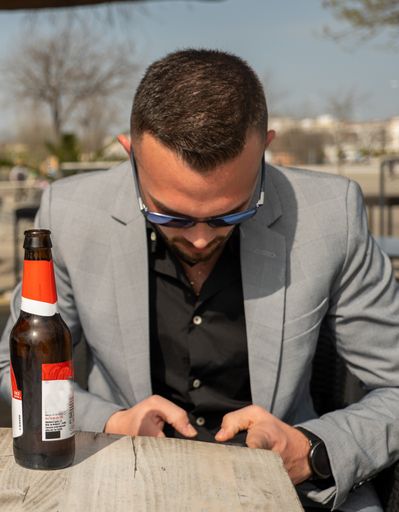 A vertical shot of a young man wearing sunglasses, a classy black shirt with a gray suit at an outdoor cafe