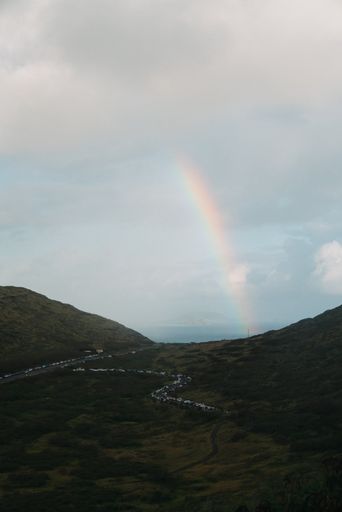A vertical shot of a rainbow in the mountain valley with a cloudy sky in the background