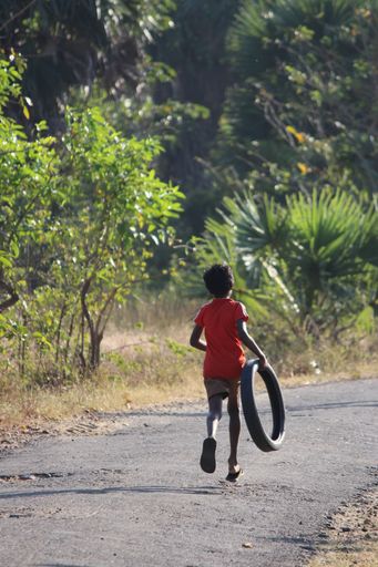 a vertical shot of a kid with a red shirt holding a black tire running down the pavement
