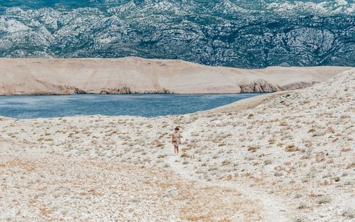 Tiny human walking on gravel path on rocky coastline. Island, sea, hiking, adventure, vast space, mountains. Island Pag, Croatia.