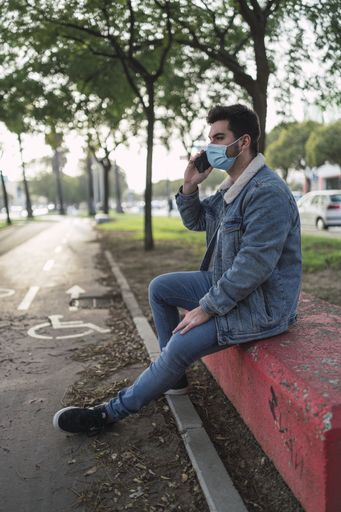 A vertical shot of a guy wearing jeans jacket, pants, a sanitary mask talking on the phone in a park