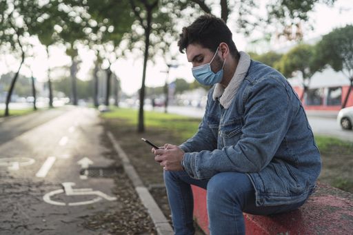 A guy wearing a jeans jacket, pants, a sanitary mask using his phone sitting in a park