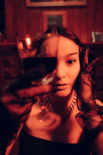 A portrait of a young sexy woman holding a glass of red wine wearing long fashionable gloves in an artificially lit room necklace