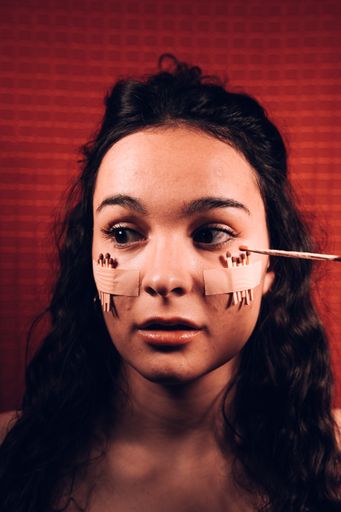 A portrait of a young woman with matches on her cheeks and a lit one in her hands on a red background