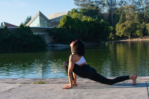 A beautiful young brunette girl doing yoga in the park, practicing Anjaneyasana pose. Doing stretching exercises with a lake in the background.