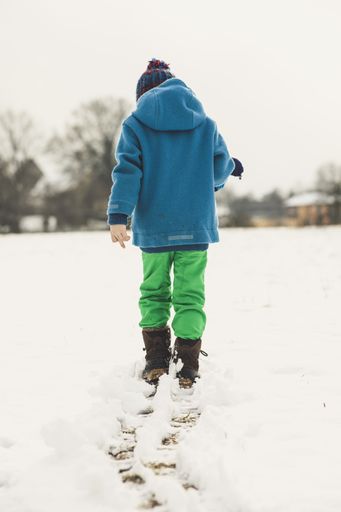 A selective focus shot of a little boy wearing a blue jacket walking in the snow from the back
