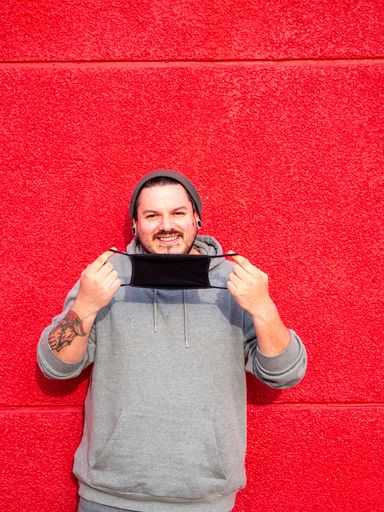 A vertical shot of a Spanish Caucasian man standing in front of a red wall, holding his facemask and laughing