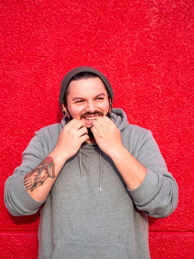 A vertical shot of a Spanish Caucasian man standing in front of a red wall, holding his facemask and laughing