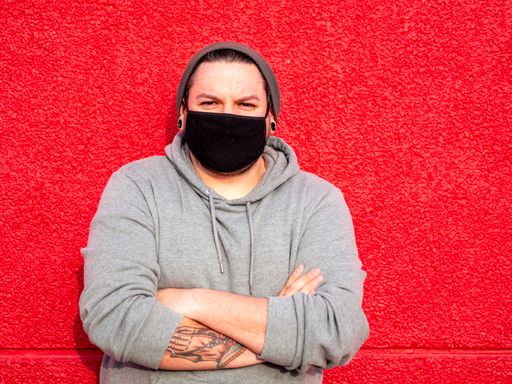 A Spanish Caucasian man standing with his arms crossed in front of a red wall, wearing a facemask