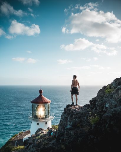 A shirtless fit male standing on a rocky cliff near a lighthouse beacon and the sea
