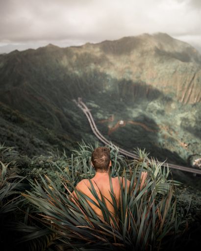 A shirtless male sitting in tall grass on a high hill with cliffs in the background