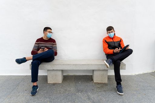 Stock photo of teenagers wearing face masks due to covid19 using tablet and talking while sitting in a bench.