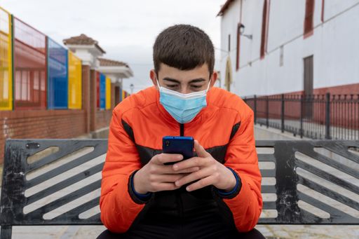 Stock photo of young boy wearing face mask due to covid19 typing on his phone while sitting in a bench in the street.