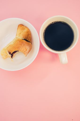 A vertical shot of a cup of coffee and bitten croissant on a white plate isolated on pink background