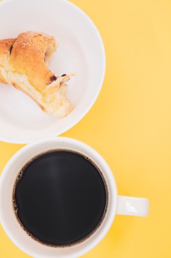 A vertical shot of acup of coffee and bitten croissant on a white plate isolated on yellow background