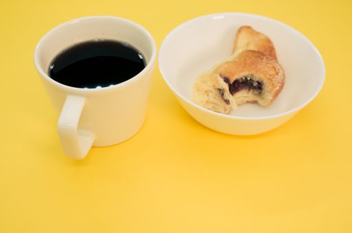 A cup of coffee and bitten croissant on a white plate isolated on yellow background