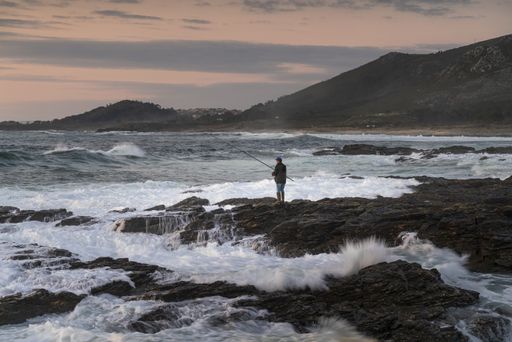 A fisherman fishing in the sea with waves crashing on the stones during sunset