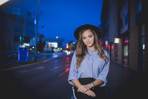 A shallow focus of an adult woman with long hair wearing a fedora hat and posing on the street at night