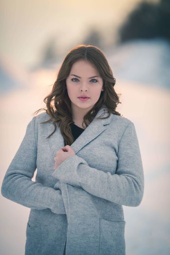 A shallow focus of a young brunette female holding her coat and posing in a field covered in the snow in winter