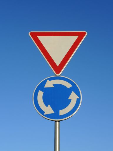 A vertical shot of two traffic signs on the same post - Give way and blue roundabout signs on the blue sky background