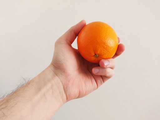 A closeup shot of a man's hand holding a ripe orange against a plain wall
