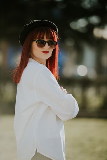A vertical side view of a young smiling female with red hair, sunglasses, and a hat posing in a park