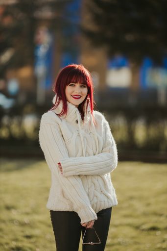 A vertical shot of a beautiful caucasian woman with red hair, wearing a white woven sweater in a park