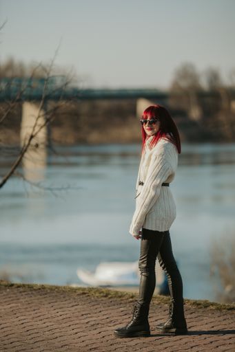 A young Caucasian woman with red hair standing on a road near a lake with a bridge in the background
