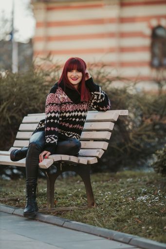 A vertical shot of a young smiling female with red hair posing in a warm sweater sitting on a bench